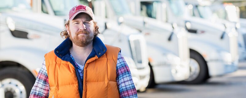 A bearded man wearing a ball cap, orange vest and plaid shirt, standing in front of semi trucks. 