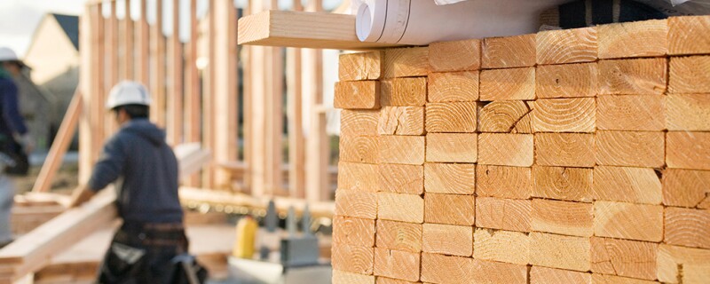 Pile of lumber on construction site with a man in the back carry pieces of wood. 