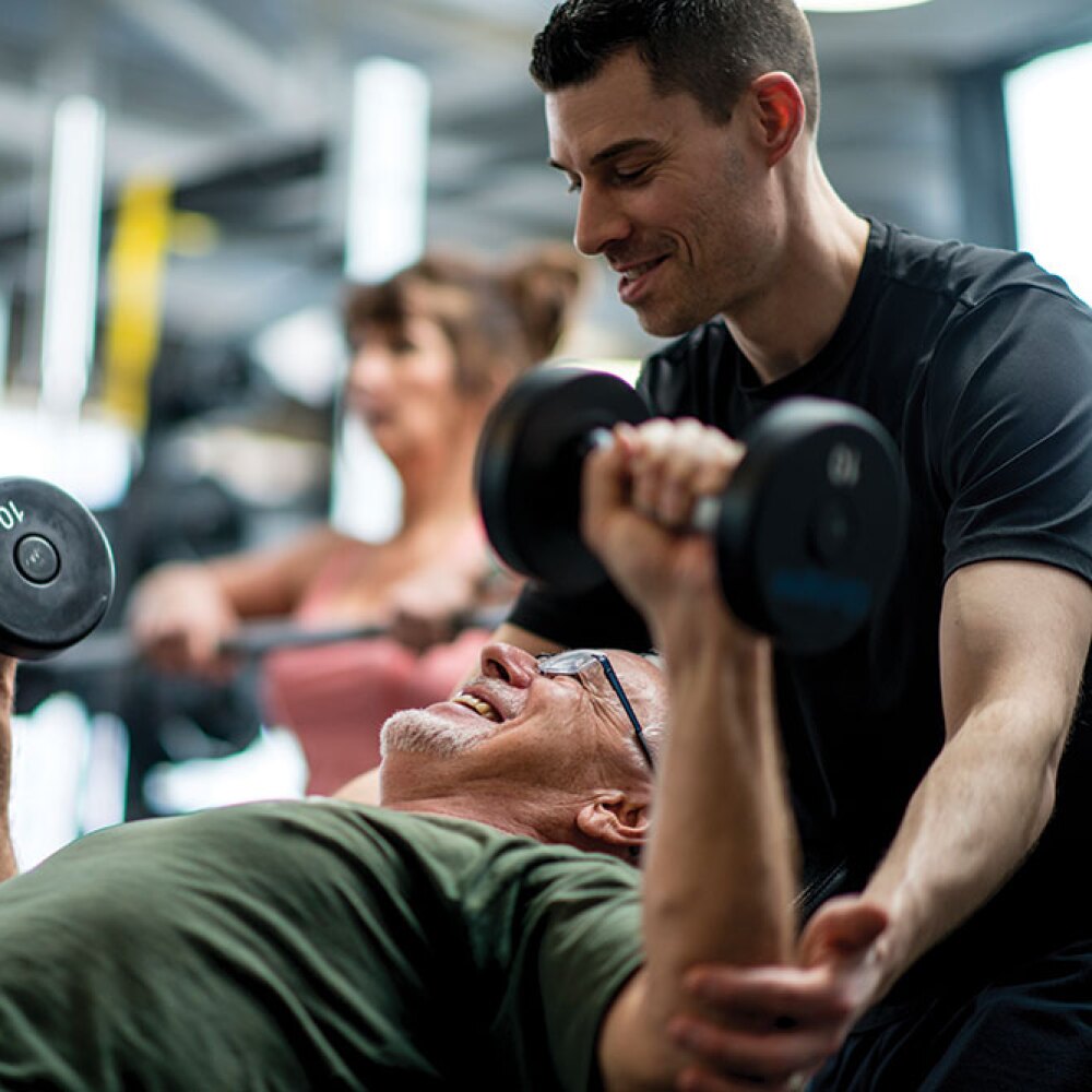 A man spotting another man that is benching free weights. 