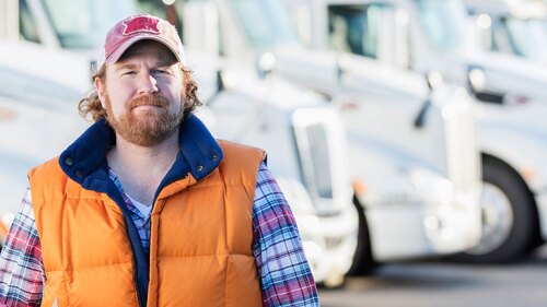 A bearded man wearing a ball cap, orange vest and plaid shirt, standing in front of semi trucks.