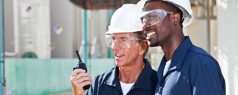 Two men with hard hats and safety glasses on with one of them talking into a walkie talkie. 