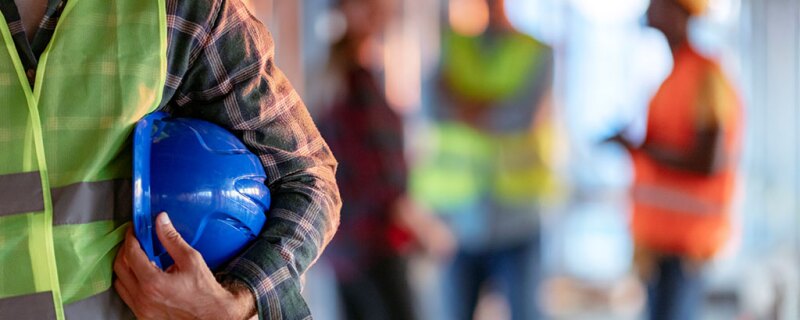 A man wearing a yellow vest and holding a blue hard hat. 