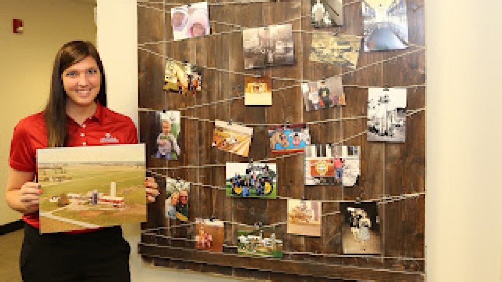 Kayla holding a photo of her families farm with other photos hanging up of her growing up on a farm.