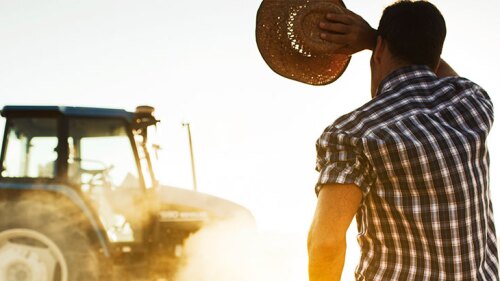 A man holding his cowboy hat while wiping his forehead and looking at his tractor.