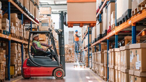 Two warehouse workers, one driving a forklift raising a pallet and the other standing on a rolling staircase taking inventory.