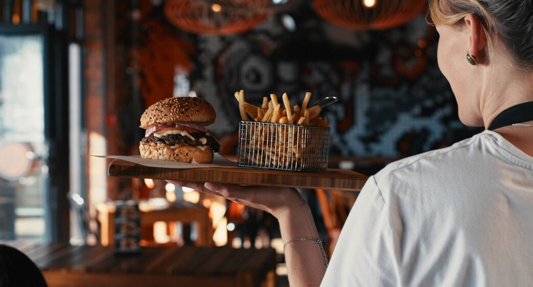 A women working as a waitress carrying a plate with a burger and fries on it to a table.