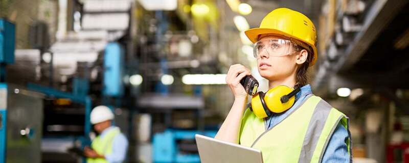 A woman wearing a hard hat, safety glasses, a yellow safety vest and has a walkie talkie in her hand.