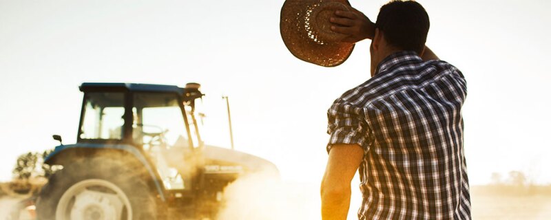 A man holding his cowboy hat while wiping his forehead and looking at his tractor. 