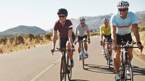 road-cycling club members riding down sunny two-lane road with mountains in the background.