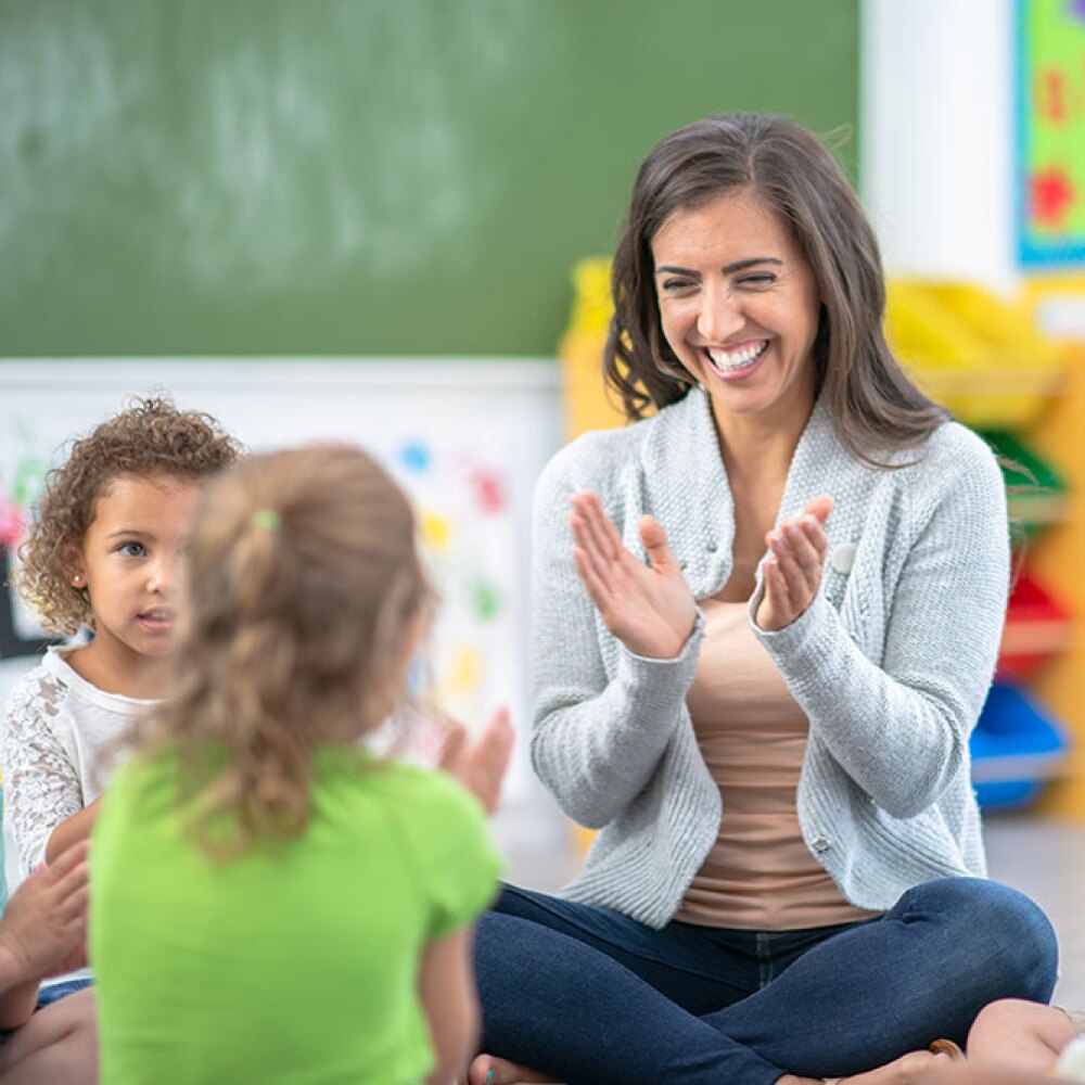 Daycare teacher sitting on the ground, smiling, and clapping with two of her students. 