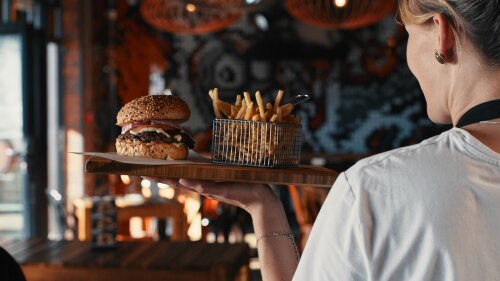 A women working as a waitress carrying a plate with a burger and fries on it to a table.