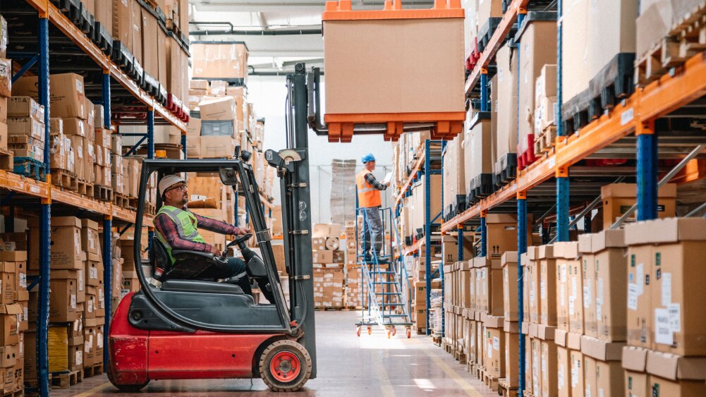 two warehouse operators, one picking up a pallet with a forklift, and the other standing on a rolling ladder inspecting inventory.